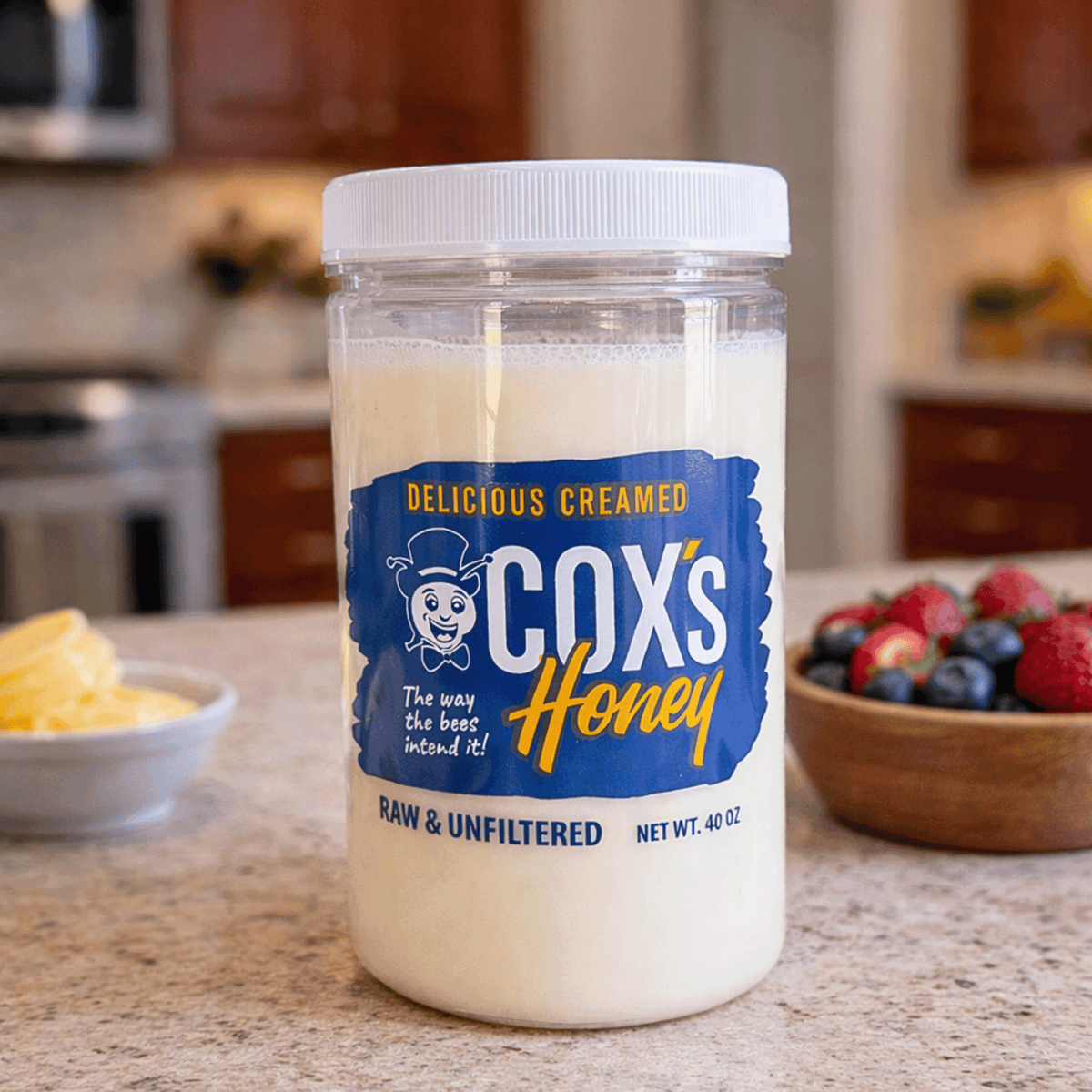 Jar of Cox's Creamed Honey on a kitchen counter with fruits in the background
