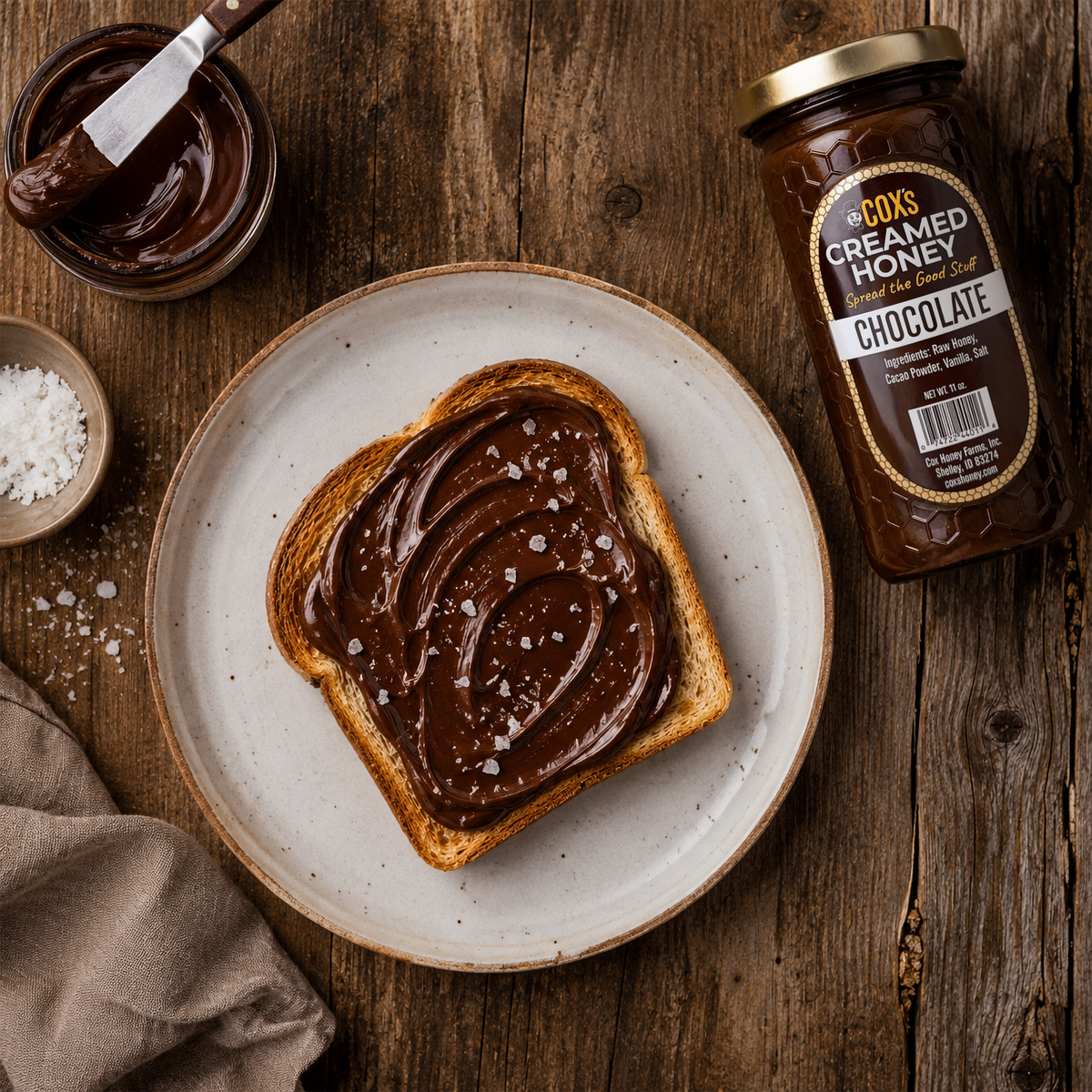 A slice of toast spread with chocolate creamed honey and coarse salt sits on a ceramic plate on a rustic wooden table, with a jar of the product and a small bowl of salt nearby.