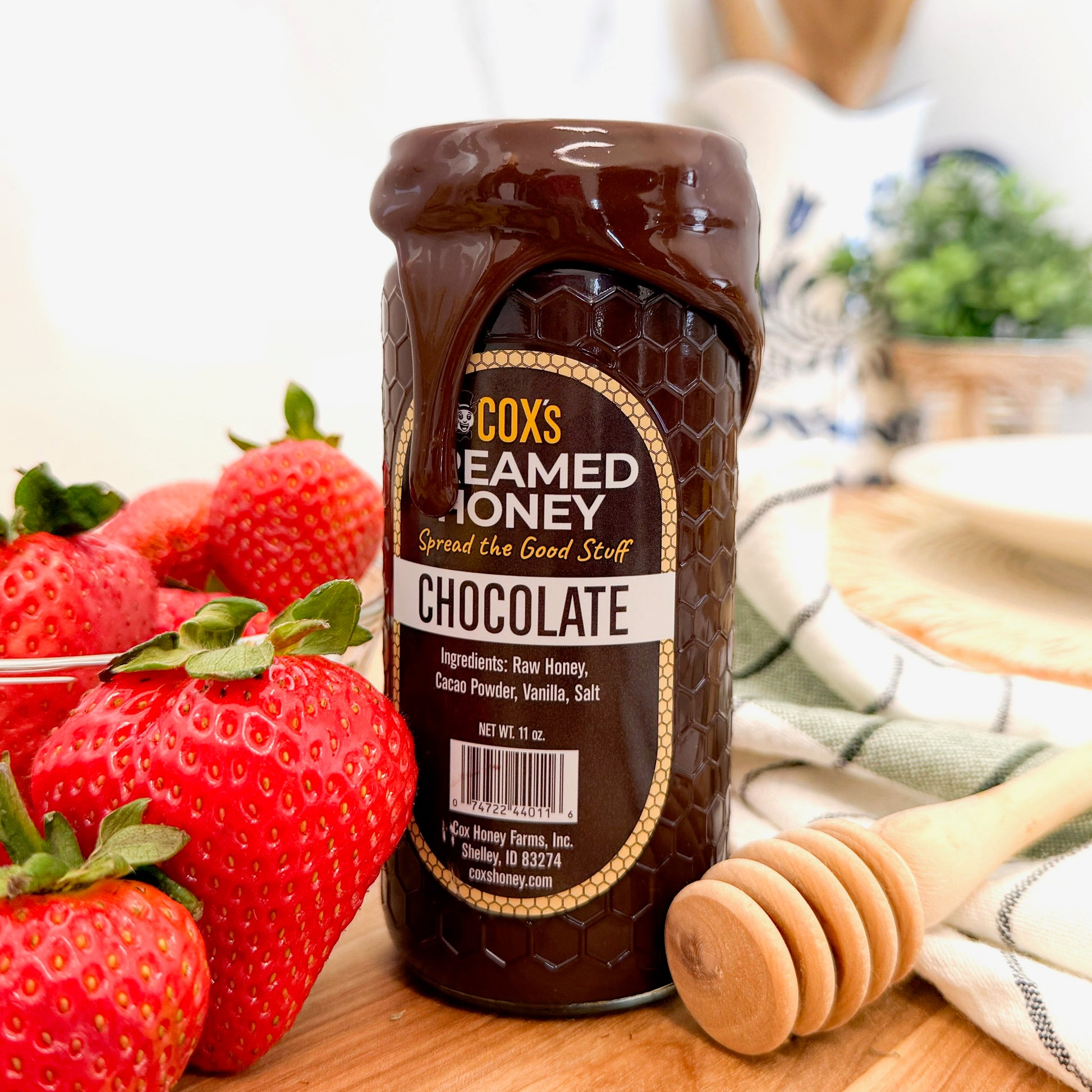 Jar of Cox's Creamed Honey Chocolate with strawberries and a honey dipper on a wooden surface.