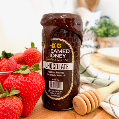 Jar of Cox's Creamed Honey Chocolate with strawberries and a honey dipper on a wooden surface.