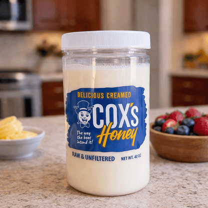 Jar of Cox's Creamed Honey on a kitchen counter with fruits in the background