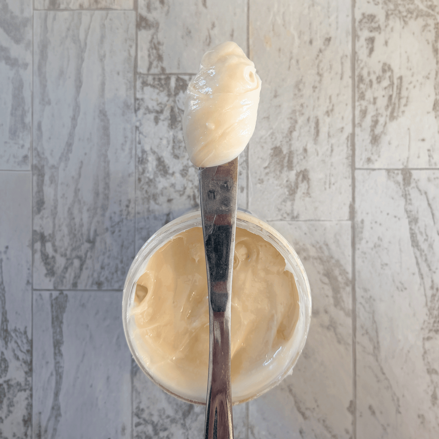 Spoon with a dollop of creamed honey over a container of cream on a marble surface
