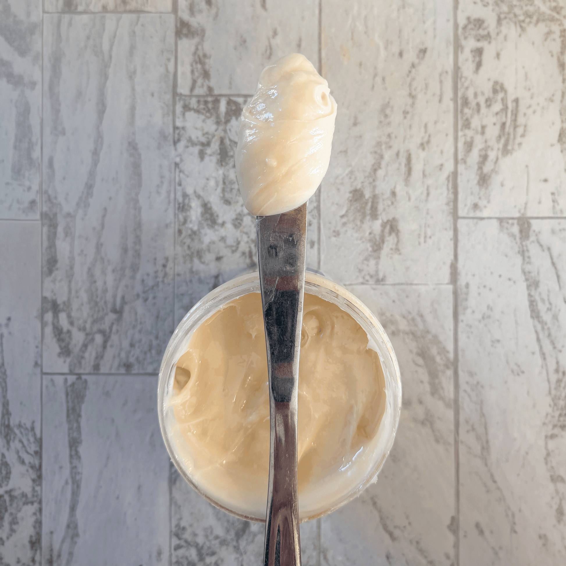 Spoon with a dollop of creamed honey over a container of cream on a marble surface