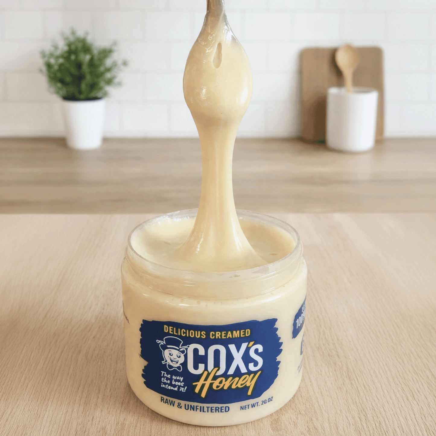 Jar of Cox's Creamed Honey with a spoon lifting honey, set on a wooden surface.