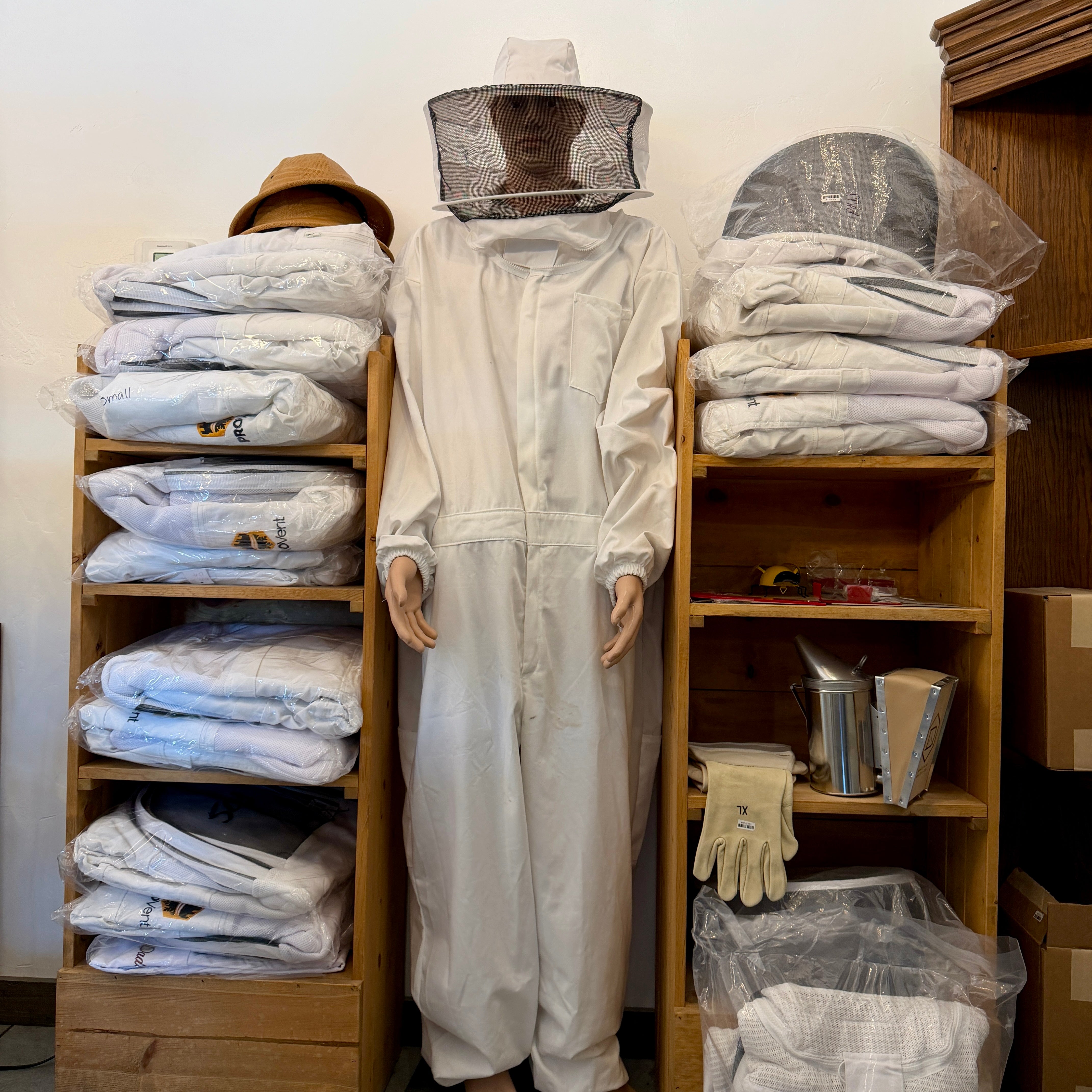 Mannequin wearing a beekeeping suit surrounded by stacked beekeeping gear in a room.