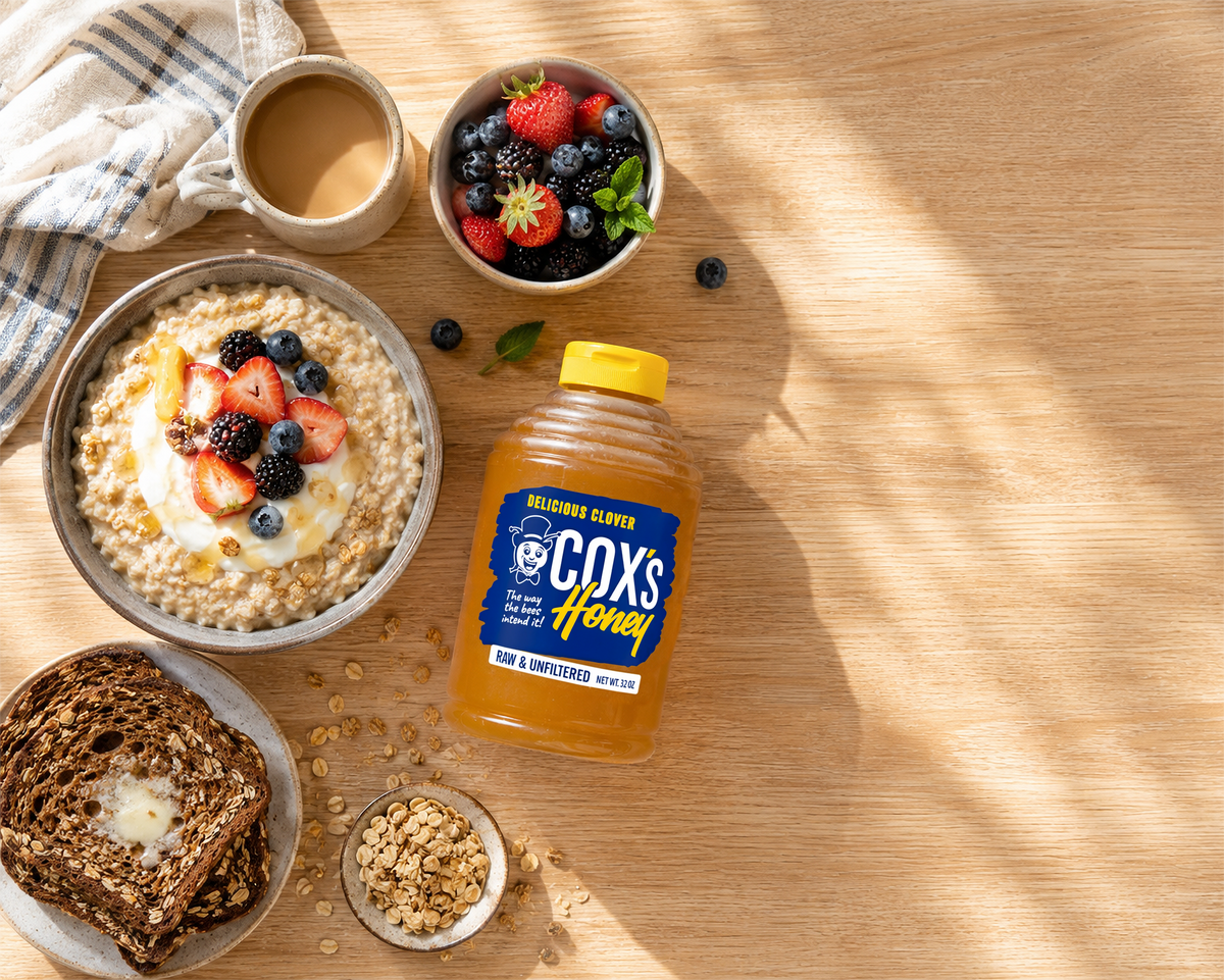 A bottle of Cox's Honey on a wooden tabletop surrounded by breakfast items including a bowl of oatmeal with fruit, yogurt, a cup of coffee, fresh berries, and slices of bread.