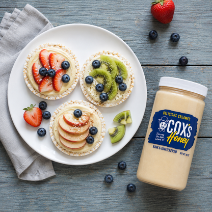 A jar of Cox's Honey Raw and Unfiltered Honey sits on a rustic blue-gray wooden table next to a white plate with three mini fruit tarts topped with strawberries, blueberries, and kiwi slices.