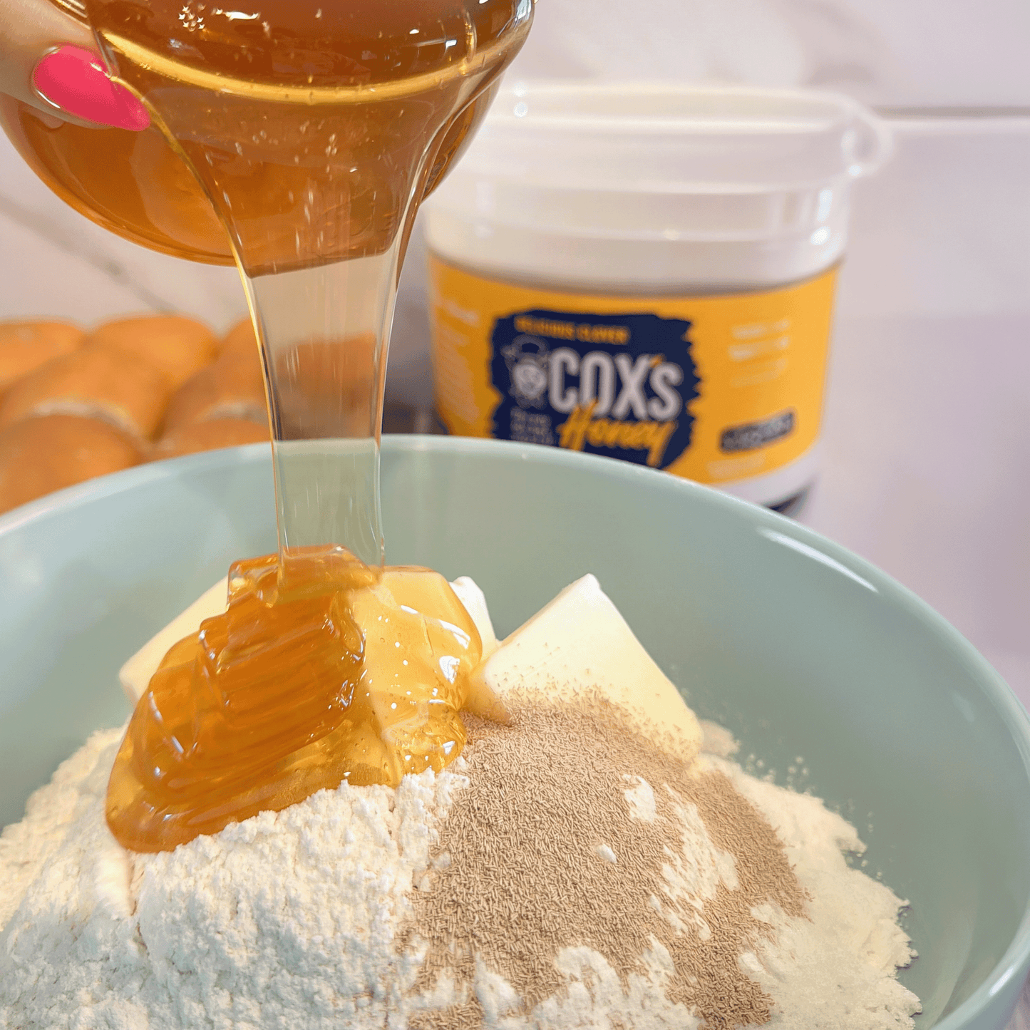 Honey being drizzled into a bowl with flour and spices, with Coxs Honey container in the background.