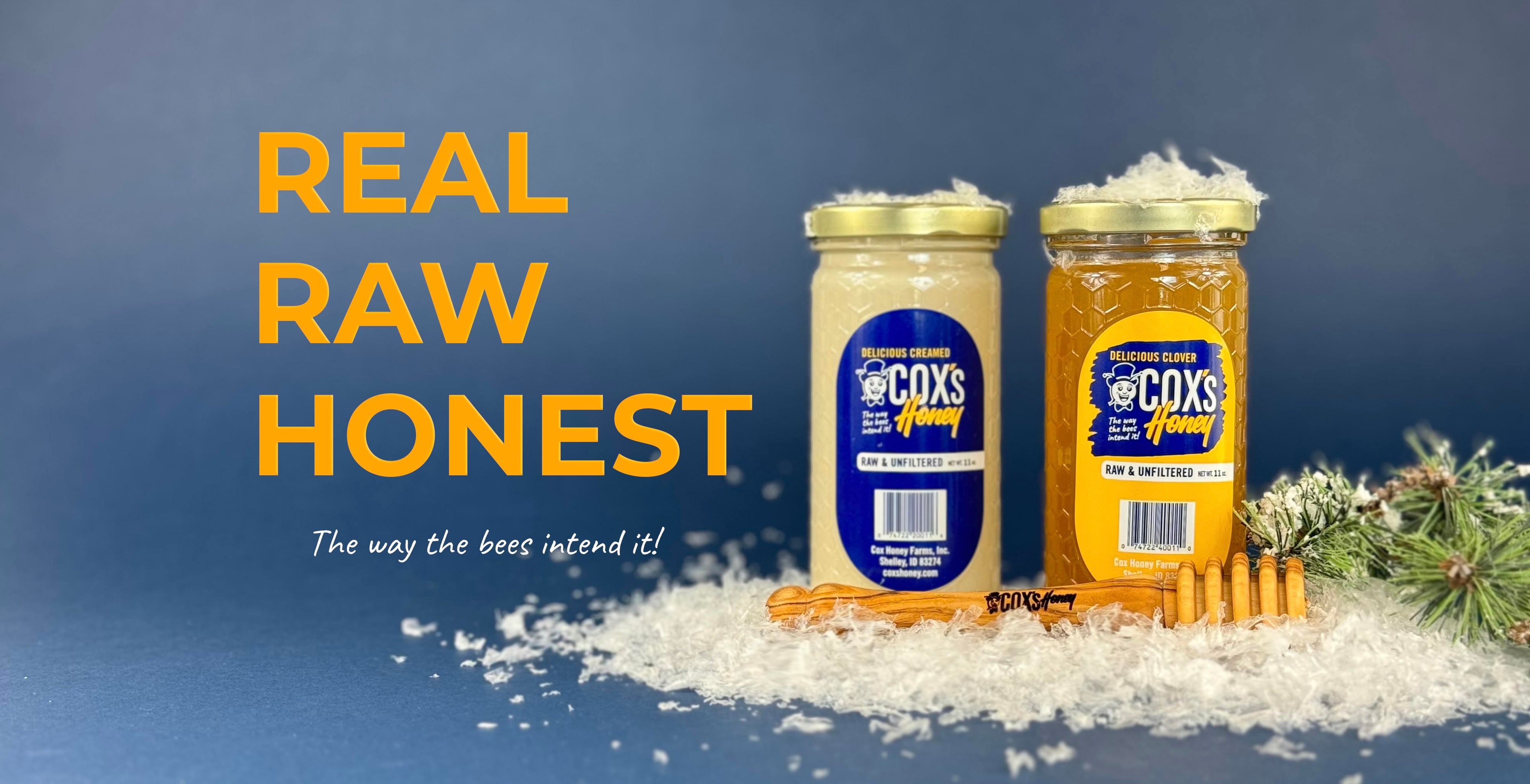 Cox's Honey Real Raw Honest commitment with The way the bees intend it slogan with a 11 oz raw, unfiltered creamed honey glass jar and a 11 oz raw, unfiltered clover honey glass jar and a olive wood honey dipper all sitting on fake snow with some green pine branches on the side