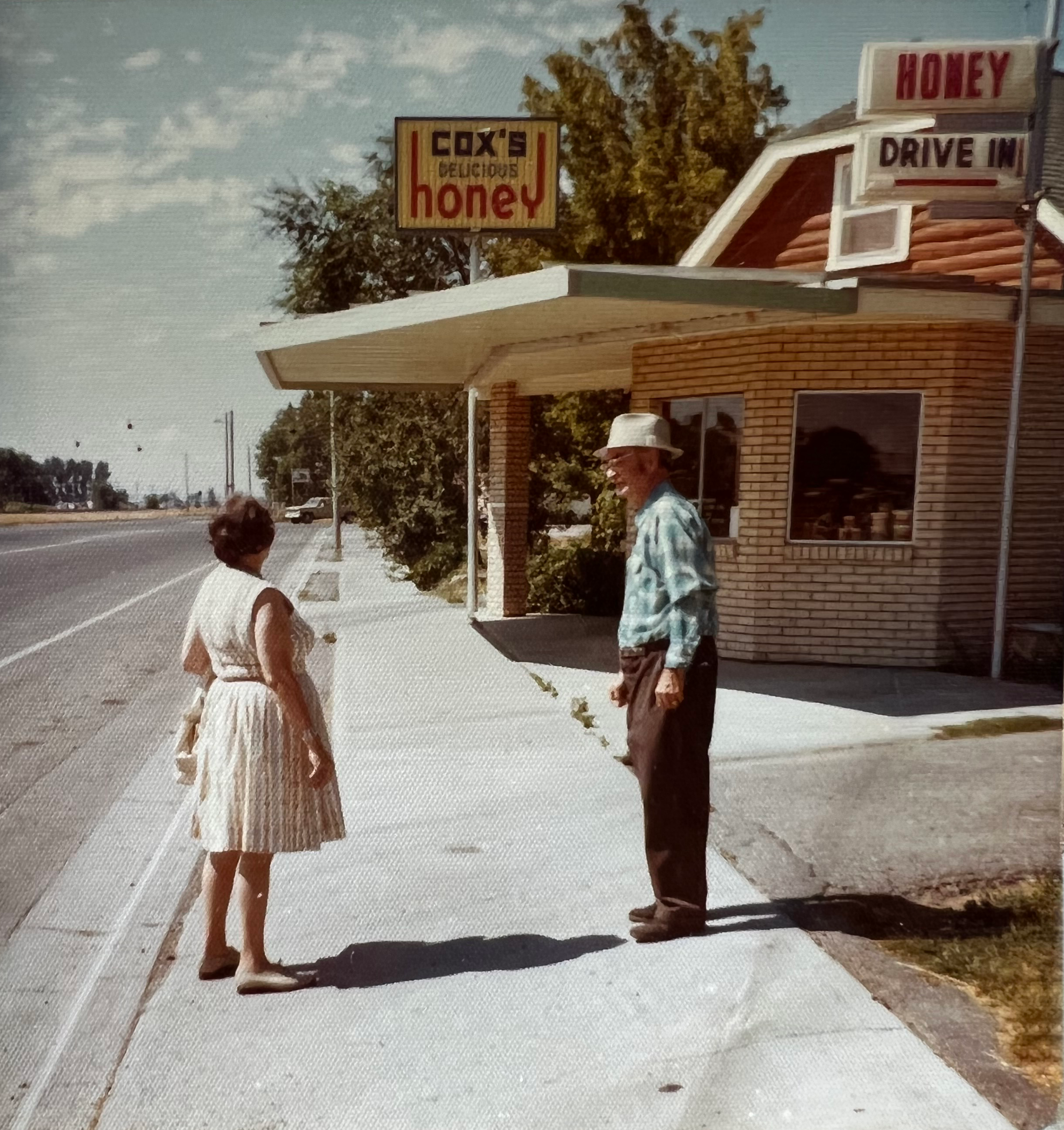 Two people standing on a sidewalk next to a small building with signs for 'Cox's Honey' and 'Drive-In'.